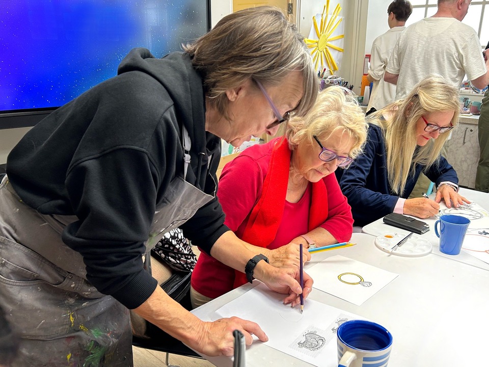 A member of staff wearing a black apron and purple glasses helping two women draw a ring.